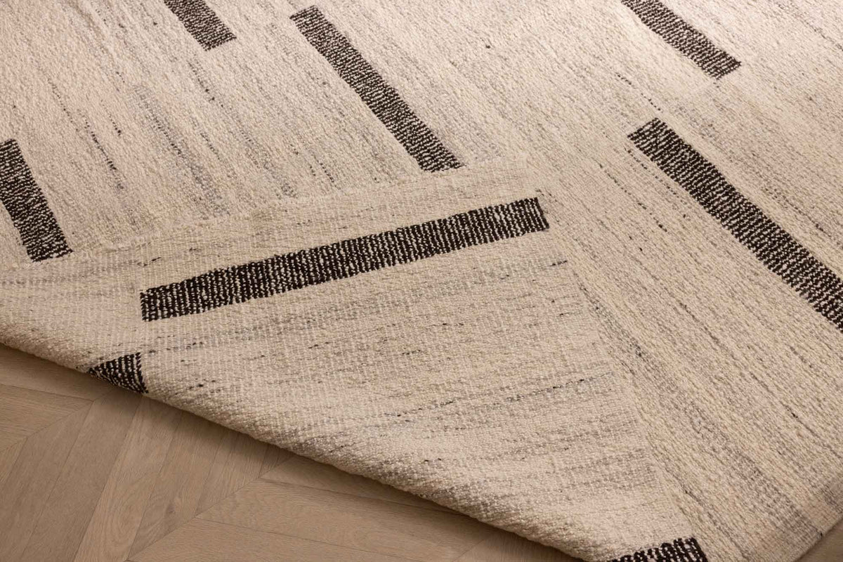 Handwoven white and brown wool-blend flatweave rug with a modern geometric stripe pattern and a folded corner.