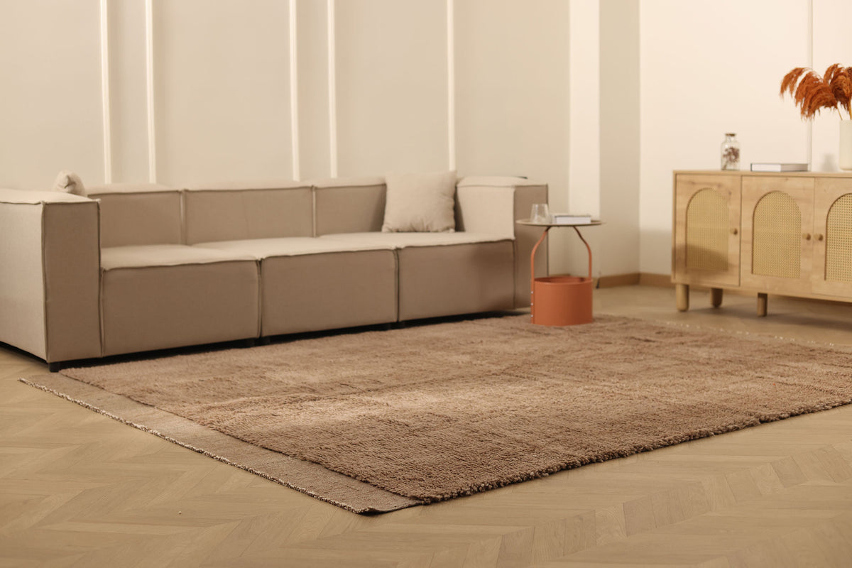 Beige sectional sofa in a living room with a brown rug and wooden cabinet.