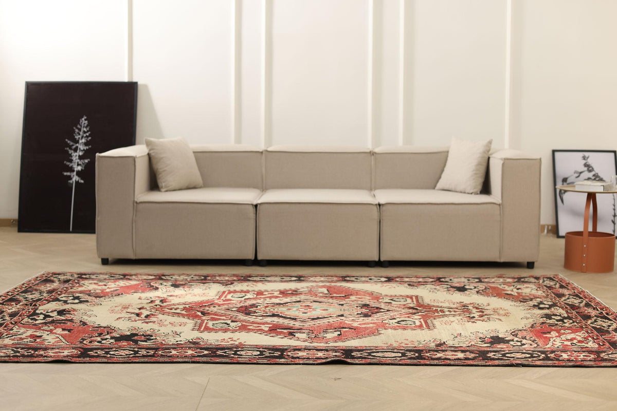 A modern sectional sofa in a light beige fabric, styled in a minimalist living room next to a large red and beige patterned oriental rug.