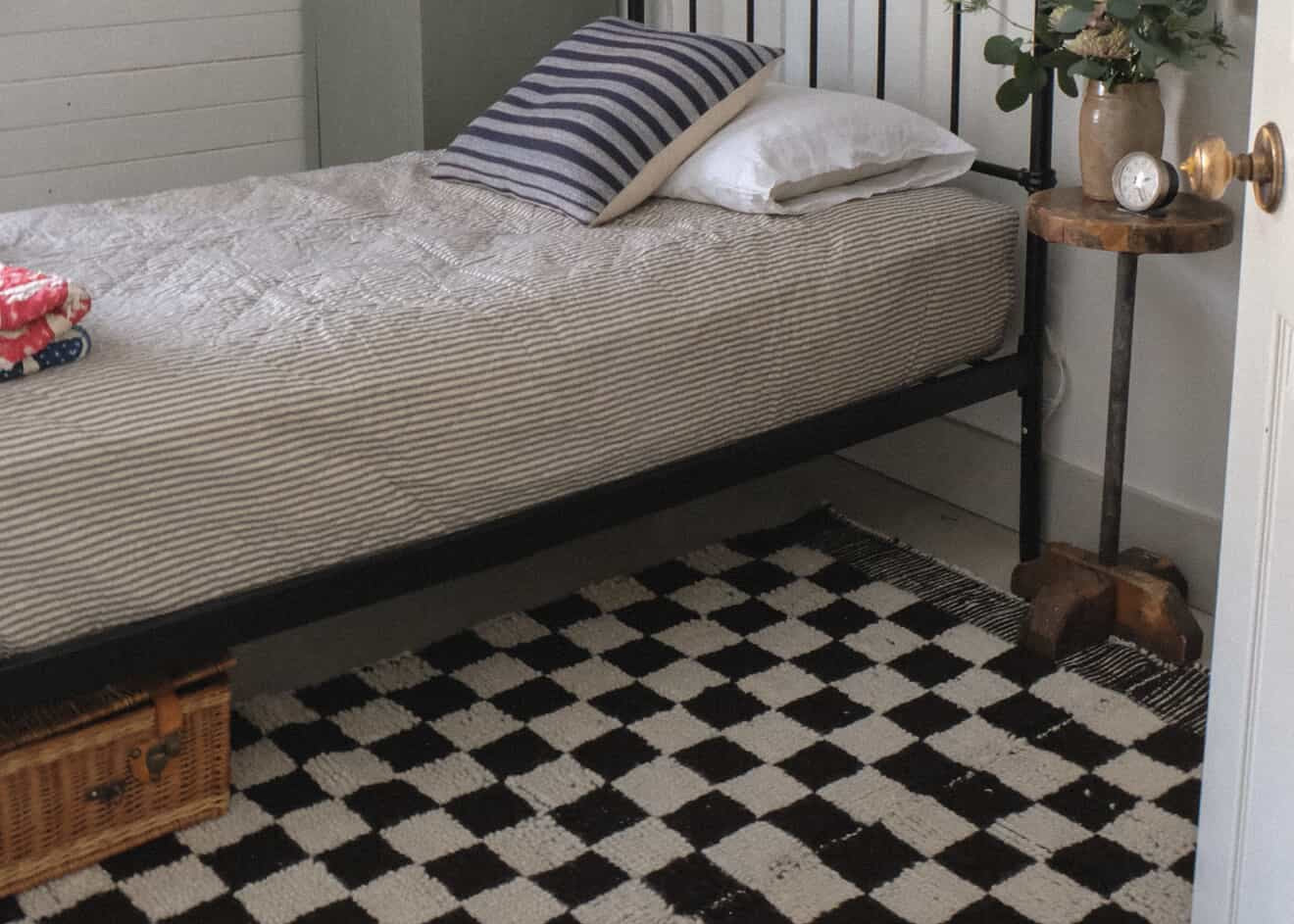A styled bedroom interior featuring a black metal single bed with a striped beige and white comforter, a striped pillow, a black-and-white checkered rug, a gray wardrobe, and framed wall art.