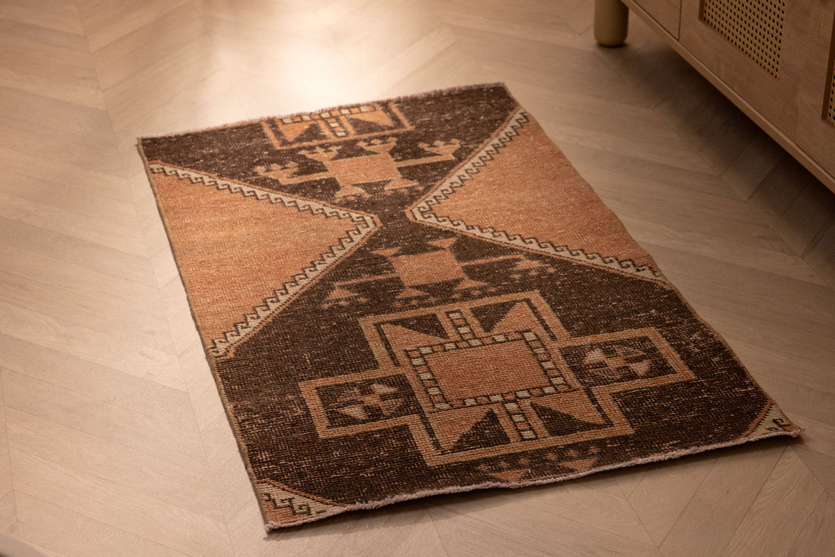 Hand-woven 2x4 ft vintage Turkish wool doormat featuring orange and brown geometric patterns on a wooden floor.
