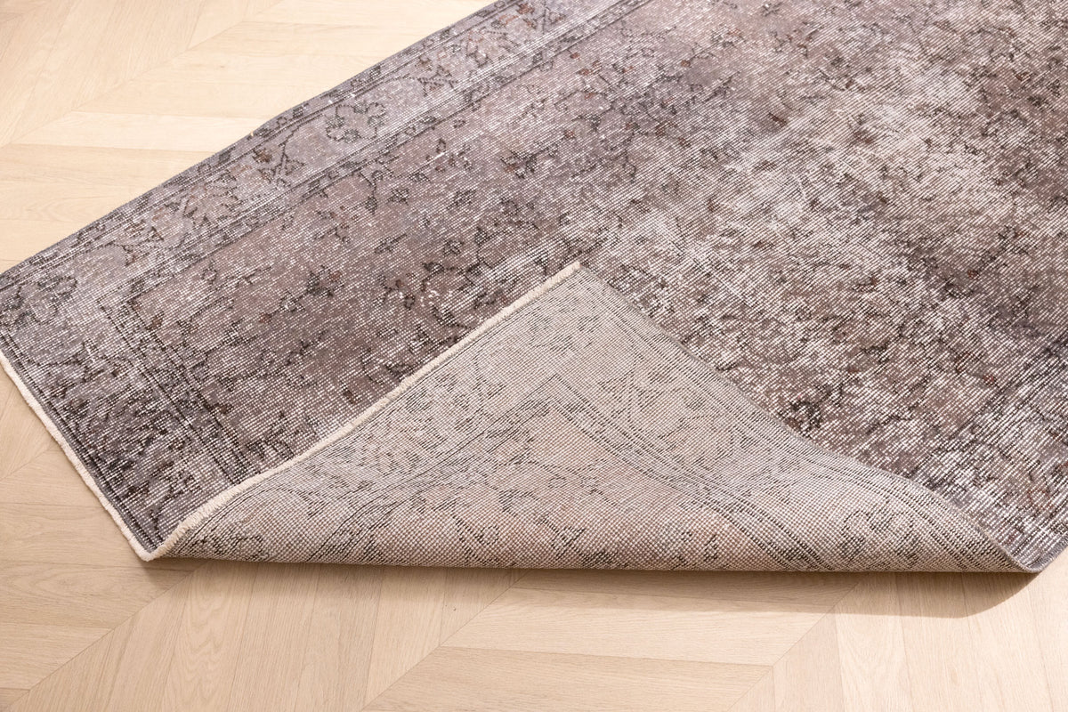 Vintage gray overdyed Isparta Turkish wool and cotton rug with floral patterns and a folded corner on a wooden floor.