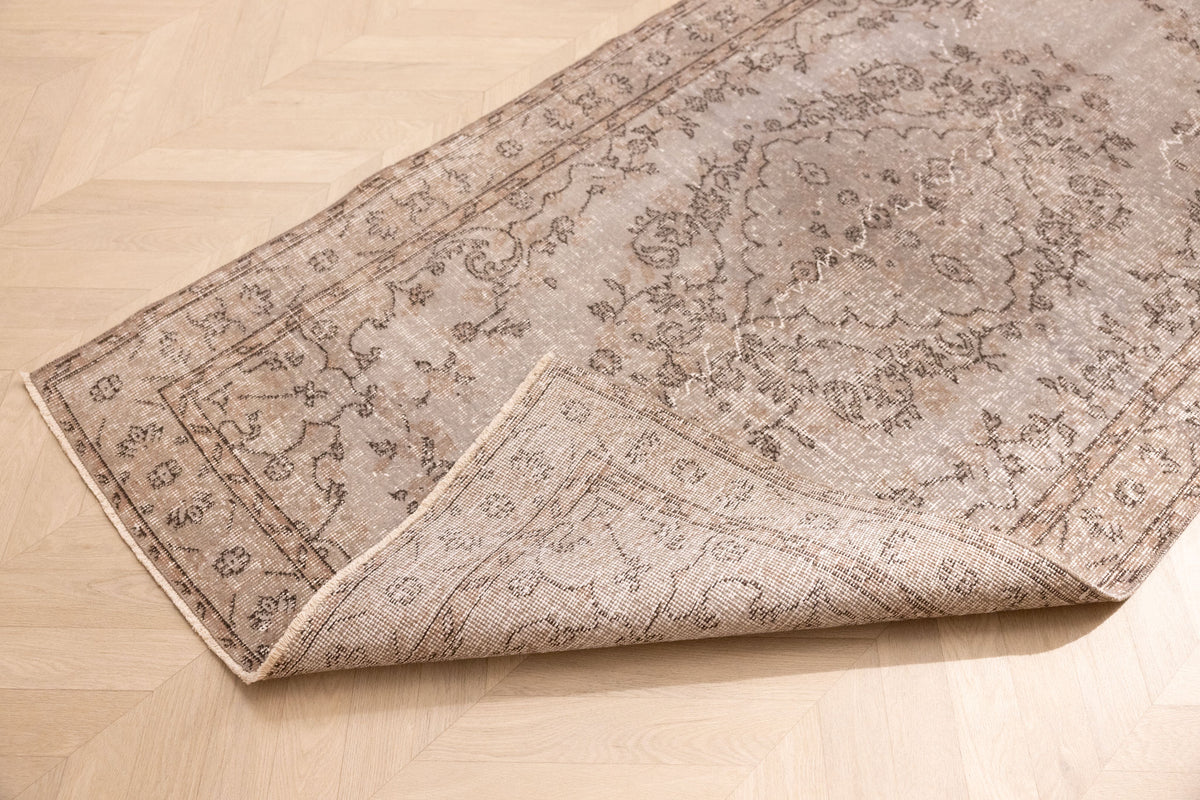 Vintage gray overdyed Isparta Turkish wool and cotton rug with floral patterns and a folded corner on a wooden floor.