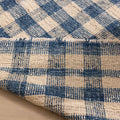 Blue and white checkered handwoven wool-blend flatweave rug with a folded corner showing texture and fringe detail.
