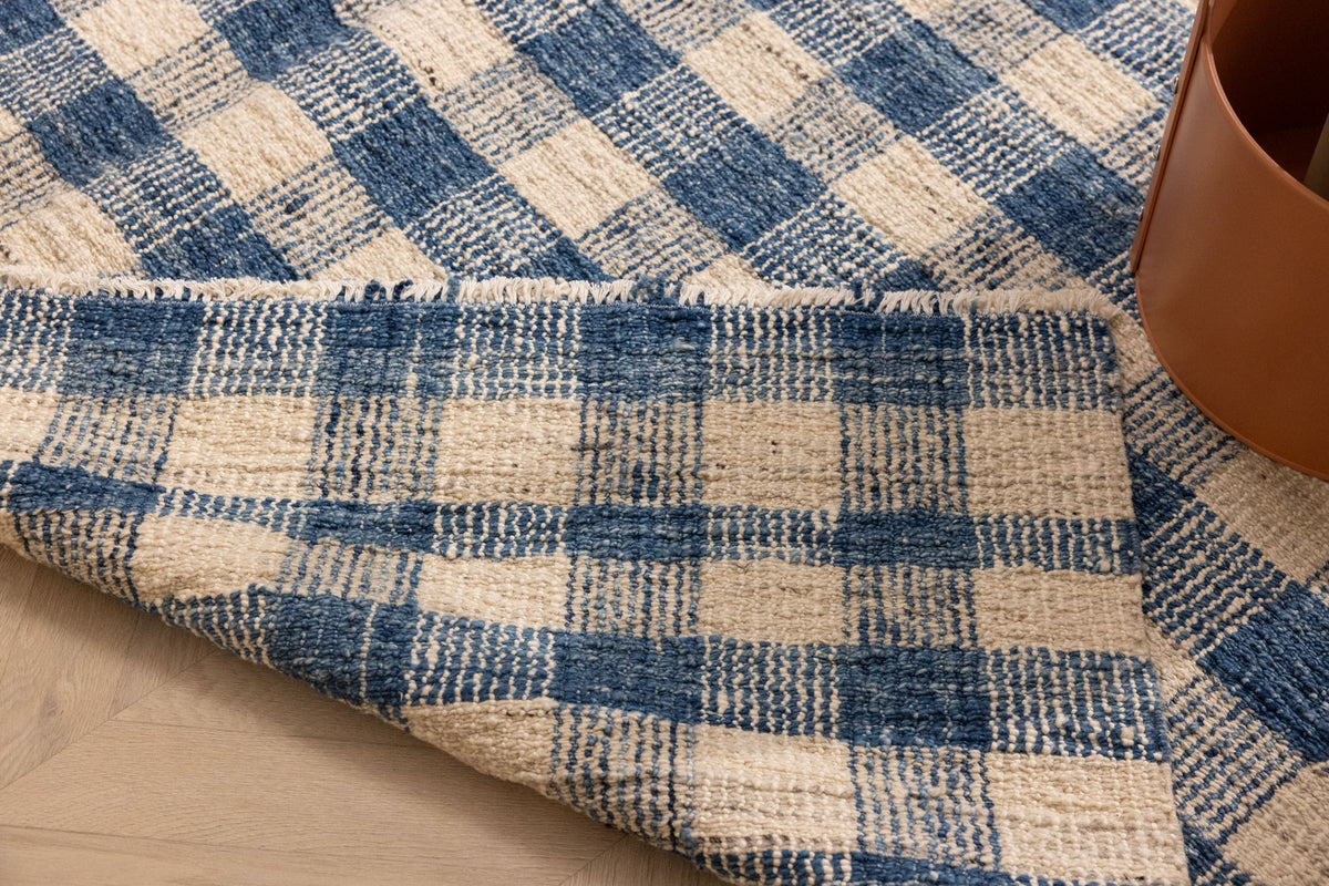 Blue and white checkered handwoven wool-blend flatweave rug with a folded corner showing texture and fringe detail.