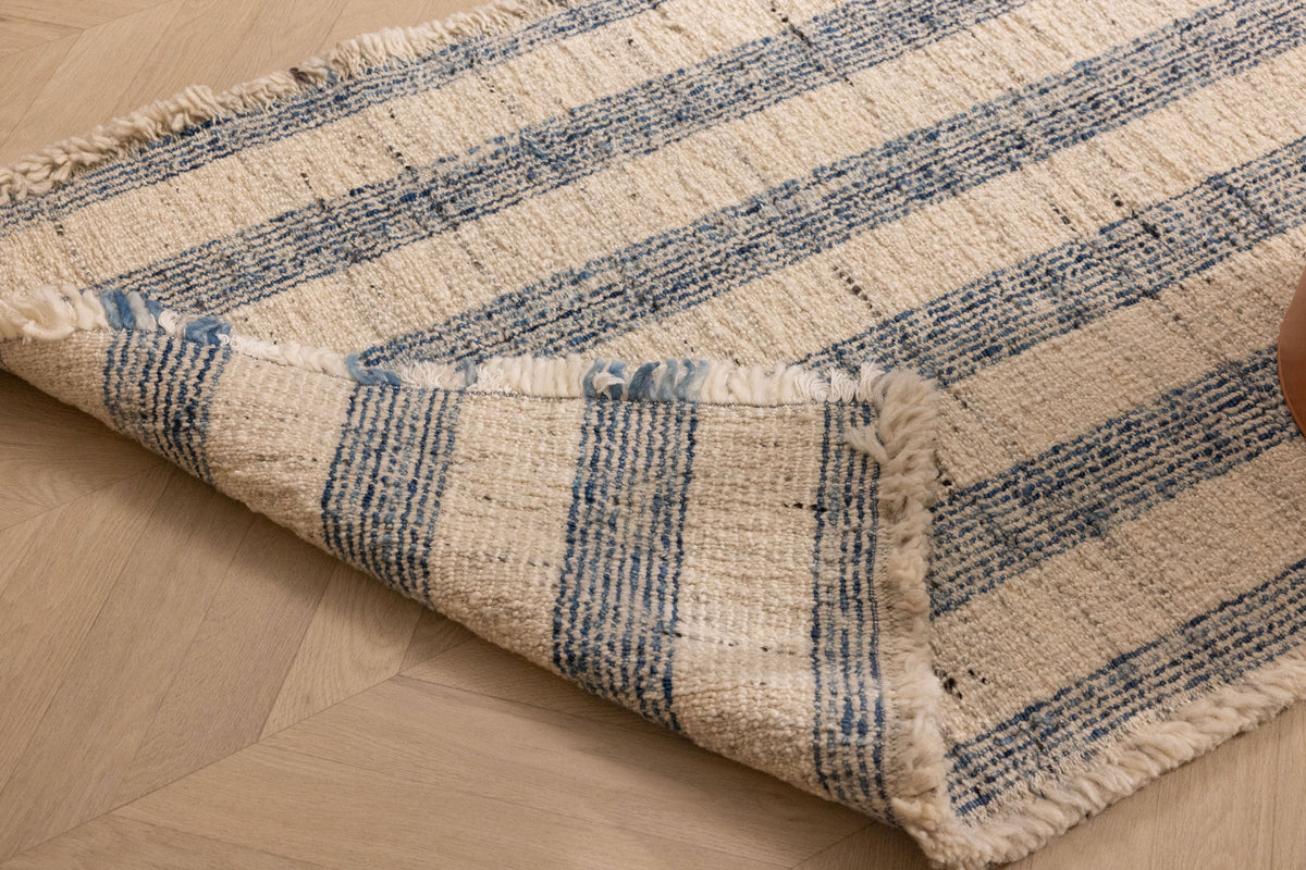 Handwoven white and blue striped wool-blend flatweave rug with decorative fringed edges on a wooden floor.