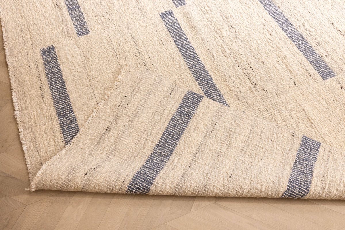 Handwoven white and blue wool-blend flatweave rug with a striped pattern, shown with a folded corner on a wooden floor.